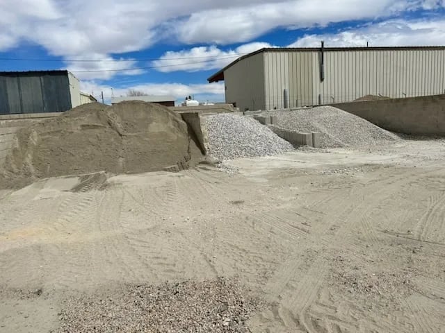 Gravel and sand piles at an industrial supply yard with metal storage buildings under blue sky