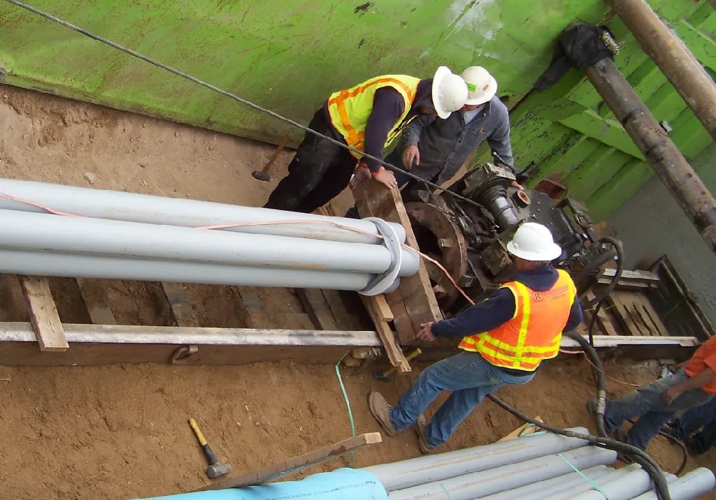 Construction workers in hard hats and safety vests laying large white PVC pipes in a dirt trench