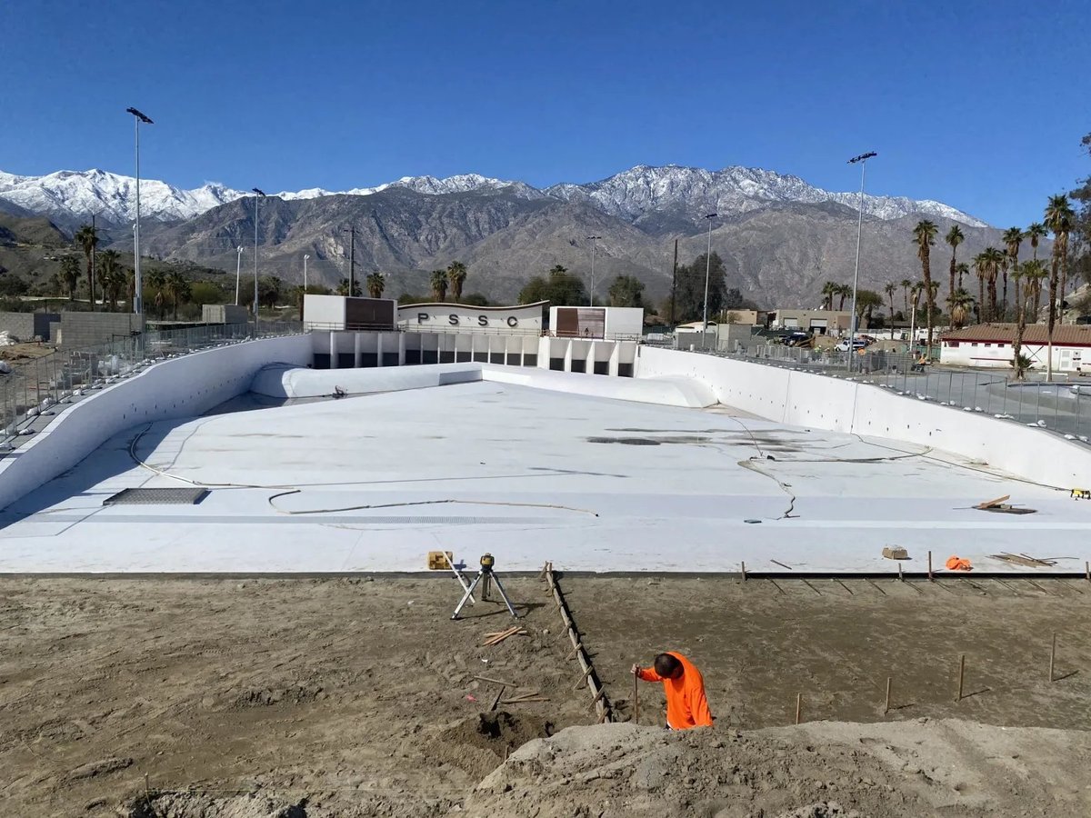 Construction site with large white roofing membrane laid out, snow-capped mountains in background, palm trees and buildings visible